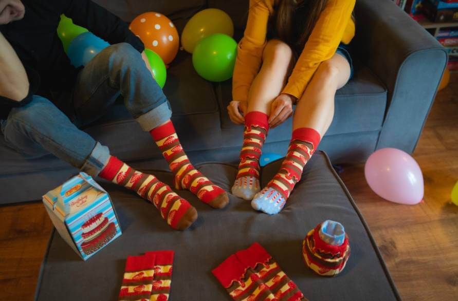 Dwie osoby na kanapie w skarpetkach Rainbow Socks z motywem tortu urodzinowego.