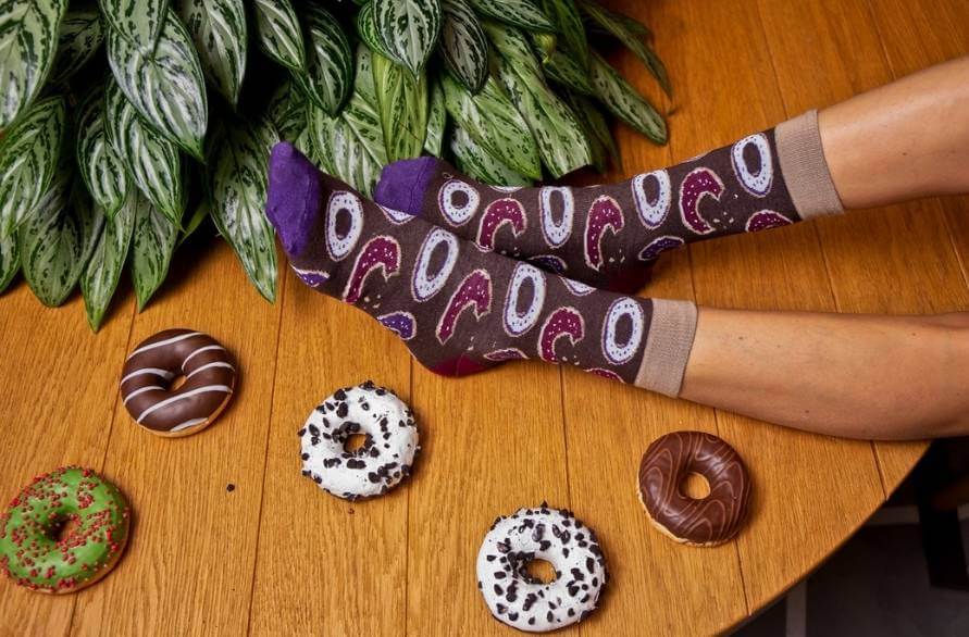 Two feet with Sweet Socks on and decorated doughnuts arranged on a wooden table.