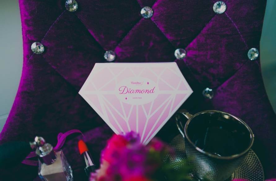 Pudełko w kształcie diamentu ze skarpetkami Rainbow Socks na fotelu glamour w otoczeniu kosmetyków.