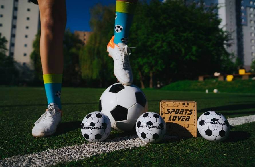 Człowiek na boisku piłkarskim z piłką i trzema zestawami piłkarskich skarpetek Rainbow Socks.