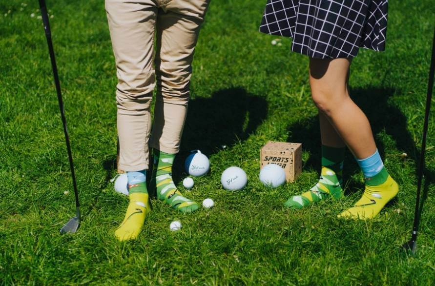 Dwoje ludzi na polu golfowym w golfowych skarpetkach Rainbow Socks w otoczeniu piłeczek golfowych.