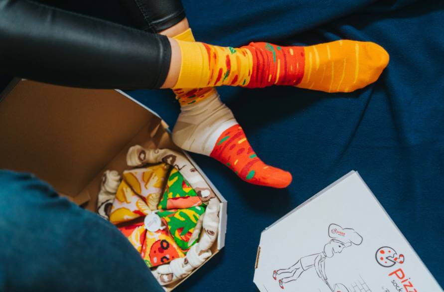 Osoba siedząca na kanapie obok pudełka z pizzowymi skarpetkami w skarpetkach pizza od Rainbow Socks.