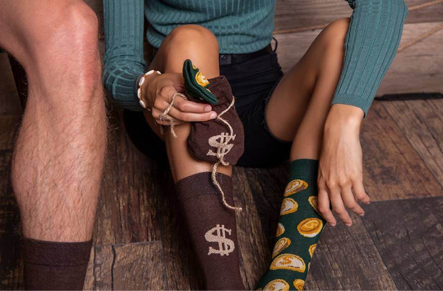 Two people in Money Rainbow socks, a woman holding a bag of money socks.