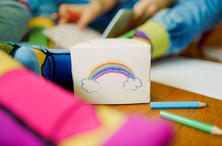 A box with a rainbow drawn with crayons next to someone’s feet in the Rainbow Crayon Socks.