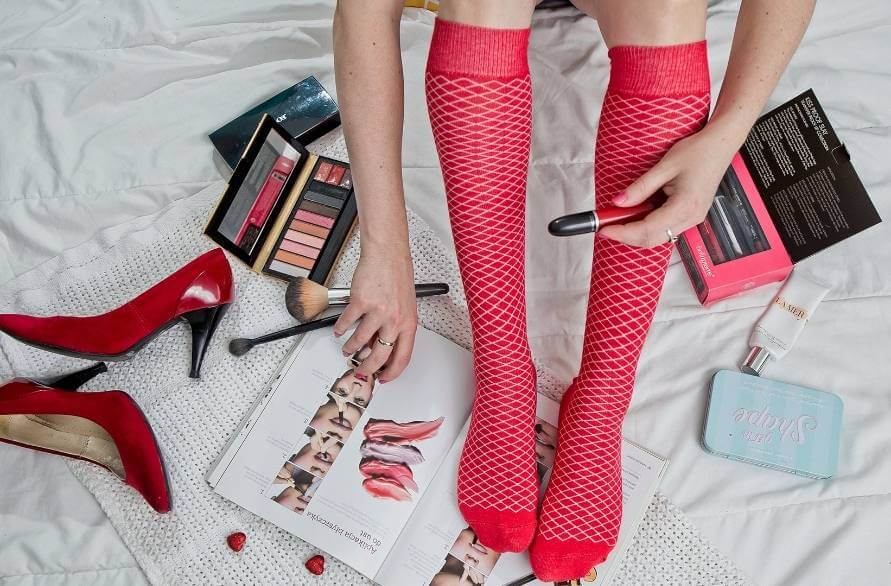 Kobiece nogi w czerwonych ażurowych podkolanówkach otoczone przez kosmetyki do makijażu.