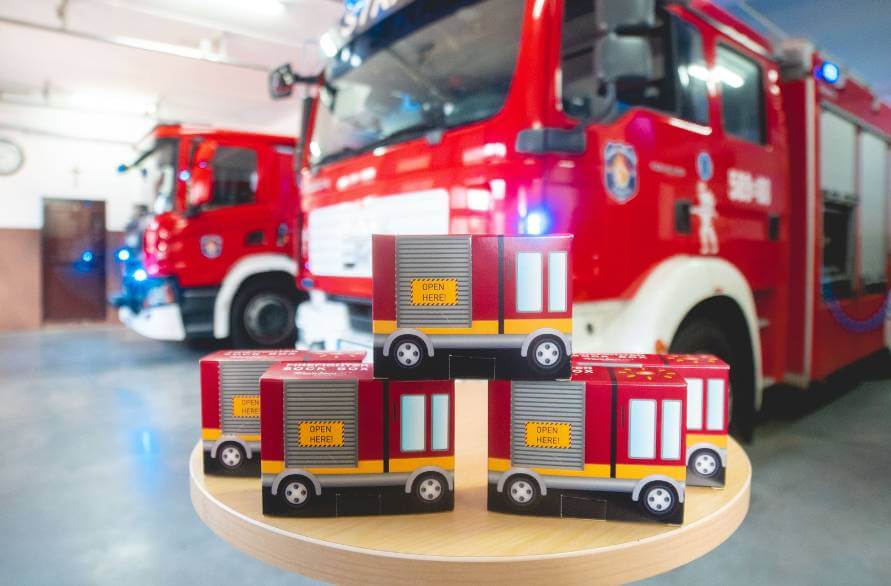 Five boxes of firefighter socks on a table with two fire trucks in the background
