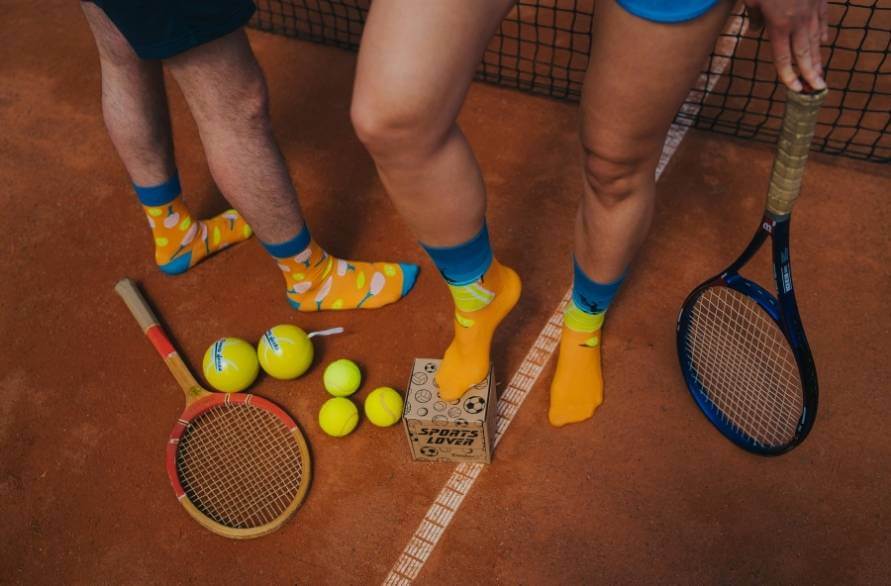 Two people on a tennis court with tennis balls and rockets, wearing the Rainbow Socks tennis socks.