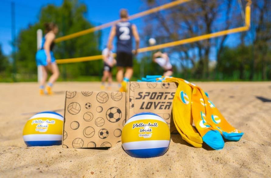 Skarpetki piłka do siatkówki od Rainbow Socks na piasku z ludźmi grającymi w piłkę plażową w tle.